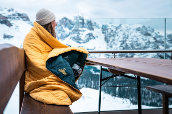 Vrouw zit buiten en kijkt uit op besneeuwde bergtoppen, gewikkeld in een warme NOMAD slaapzak