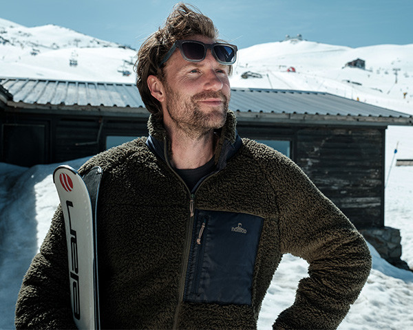 Een man met een Sherpa fleecvest aan en ski's in zijn handen bij een houten huisje met sneeuw.